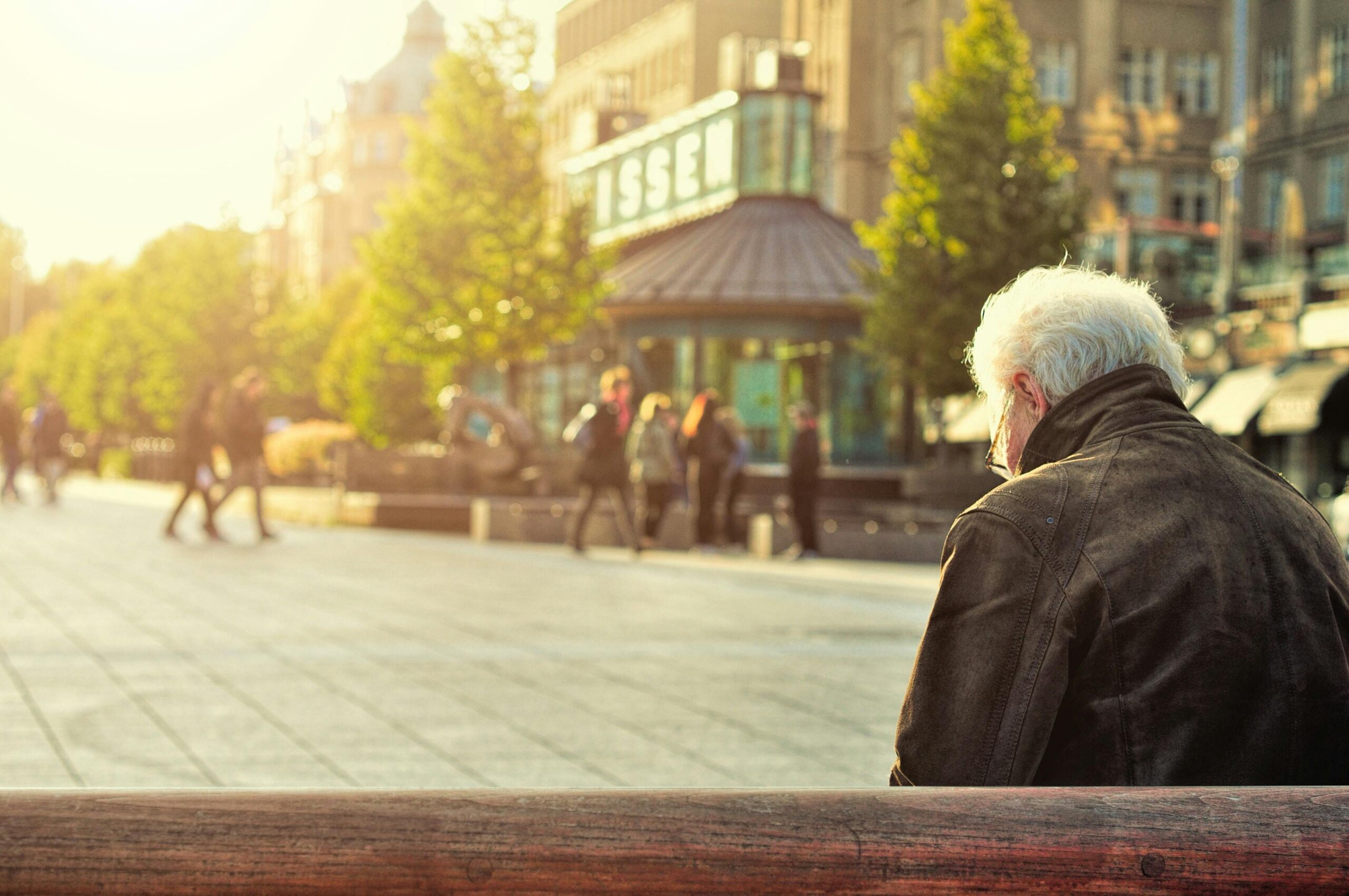Homem idoso senta em banco olhando para a cidade, representando investimentos em saúde e longevidade.